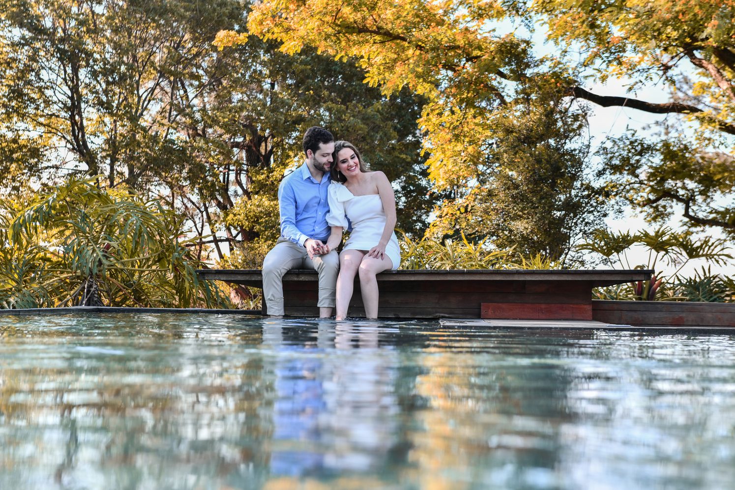 Casal em ensaio sentado à beira da piscina com árvores douradas ao fundo