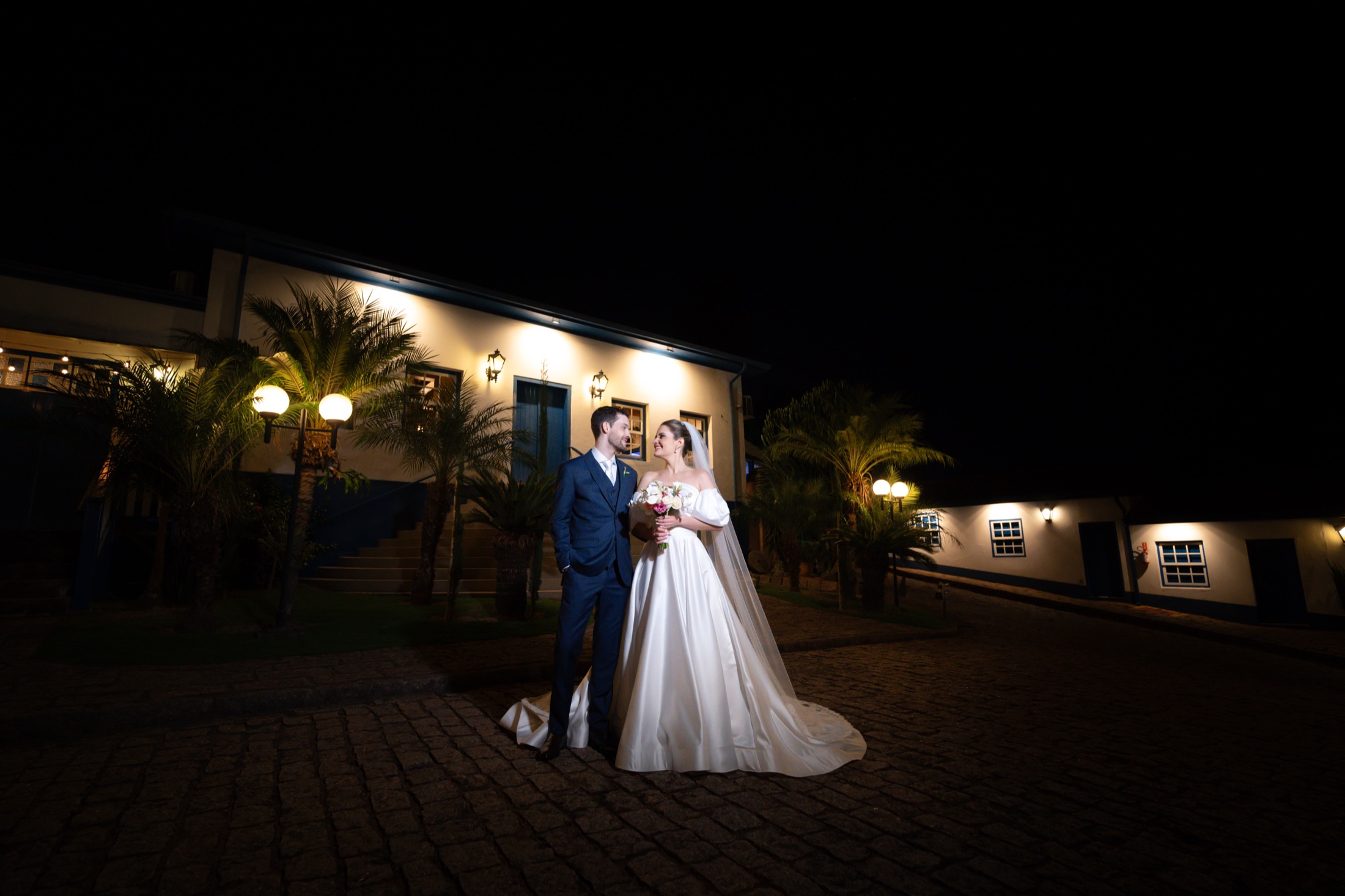 Casal de noivos em retrato noturno diante de casa de festas iluminada, fotografado pelo Studio9